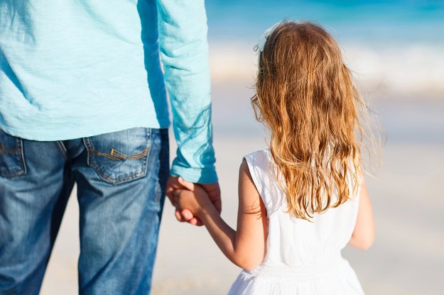 A young girl holding her father's hand at the beach.