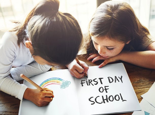 Two girls in school coloring a rainbow and writing: First Day of School.