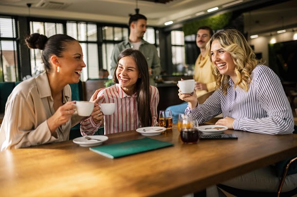 Three women laughing while sitting in a cafe with coffee cups in their hands.