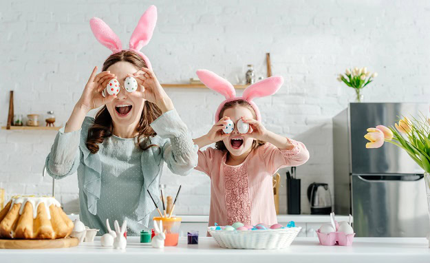 A happy mom and daughter in the kitchen holding up Easter eggs in front of their eyes.