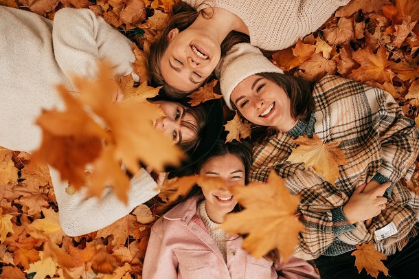 Women lying on autumn leaves and smiling while more leaves fall on them.