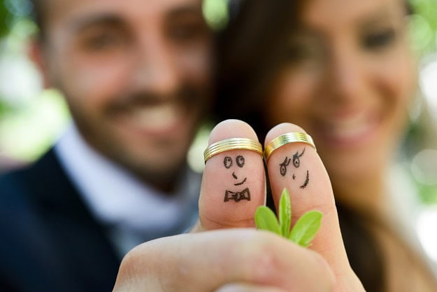 A newly wed couple with their thumbs up with smiling faces drawn on them and the wedding rings as thumb crowns.