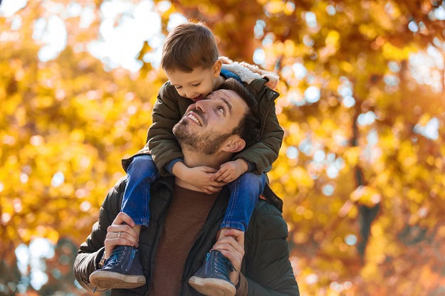 A smiling son riding on his father's shoulders in a fall landscape with leaves of all colors.