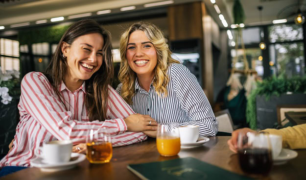Two happy women smiling into the camera while at a coffee shop.
