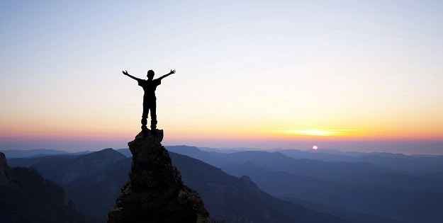 A man at a top of a mountain stretching his hands upward in celebration during sunset.