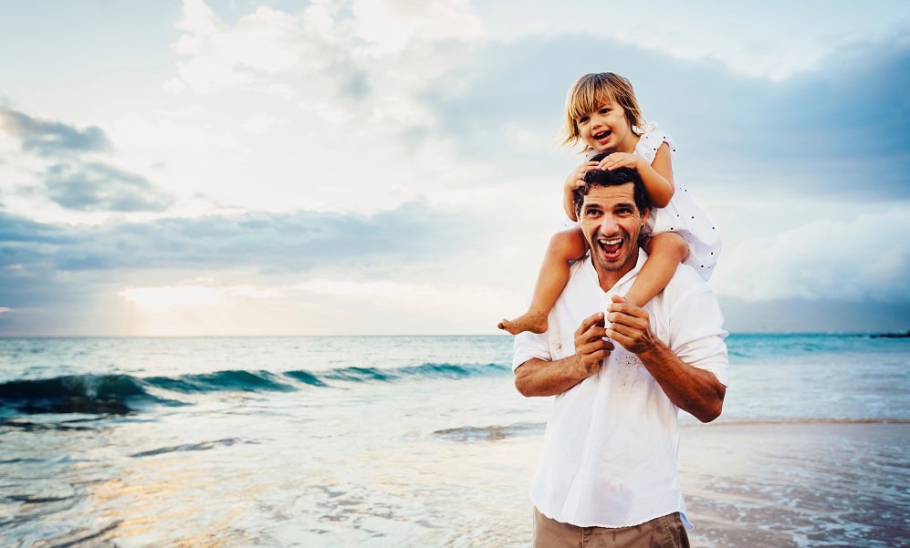 A happy dad with a his daughter on his shoulders at the beach.
