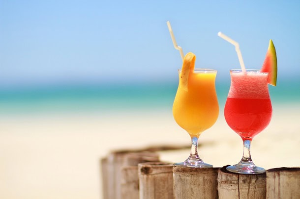 Two colorful juice drinks with straws and a sandy beach and blue ocean in the background.