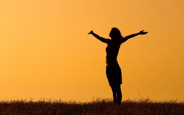 A woman at sunrise outside stretching her arms towards the sky.,