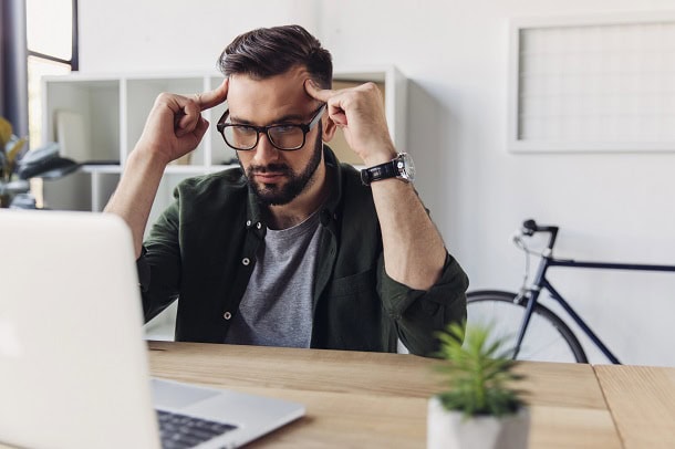 A man sitting by his laptop, thinking and looking frustrated.